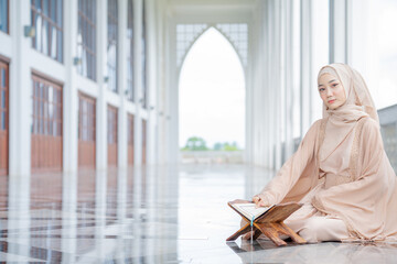 The image of an Asian Muslim woman in the Islamic religion in hijab in white color. reading the Quran, Koran Staying in a beautiful mosque, Arabic word translation: The Holy Al Quran.