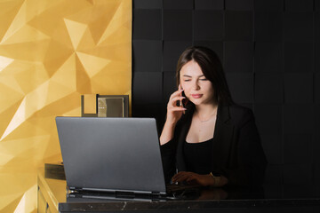 A young woman is talking on a smartphone at a table in front of a laptop