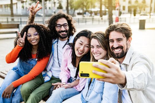 Diverse group of friends taking a selfie in the street sitting on a bench. Cheerful multiracial group of young hipsters taking a picture outside in a park bench.