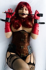 Red-haired woman in a red wig, corset and leather gloves posing lying on a bed holding a stick in...
