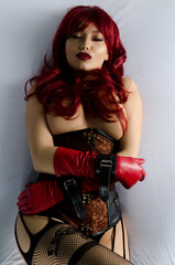 Red-haired woman in a red wig, corset and leather gloves posing seductively lying on the bed