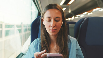 Cute girl with long brown hair, wearing blue shirt with smartphone and wearing wireless earphone into ear traveling by suburban train