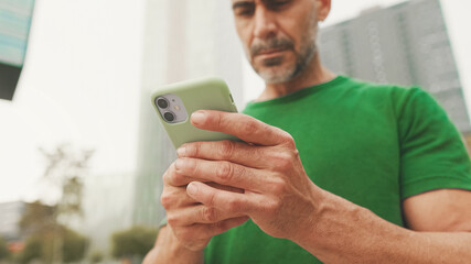 Mature man wearing casual clothes, standing on modern buildings background, texting on smartphone
