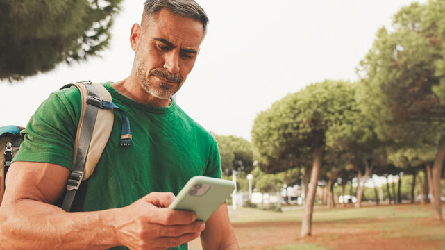 Mature man wearing casual clothes, with backpack on his shoulder, stands in the park, sends SMS from mobile phone, always online
