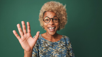 Close-up of a smiling girl in glasses wearing a blue t-shirt, looking at the camera, taking a selfie, waving her hand in greeting, isolated on green-blue background