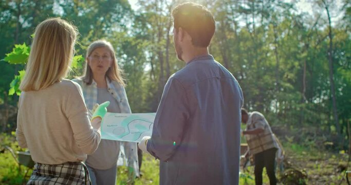 Male and female ecological activists standing with map or plan in hands and giving commands to people for planting trees in forest outdoors. Enviromental concept. Charity work of volunteers.