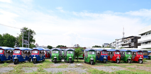 BANGKOK, THAILAND - JULY 10, 2023: Several Electric tuk tuks taxi stopped in the meadow with buildings background. Concept to save fuel, maintain and reduce air pollution protect the environment.