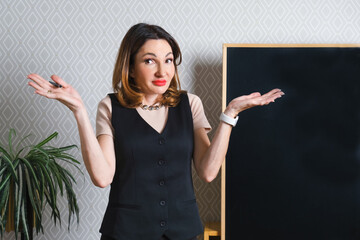 Attractive middle-aged woman in a black vest stands by an empty slate board. Woman spread her hands in disbelief. Space for text on the board.