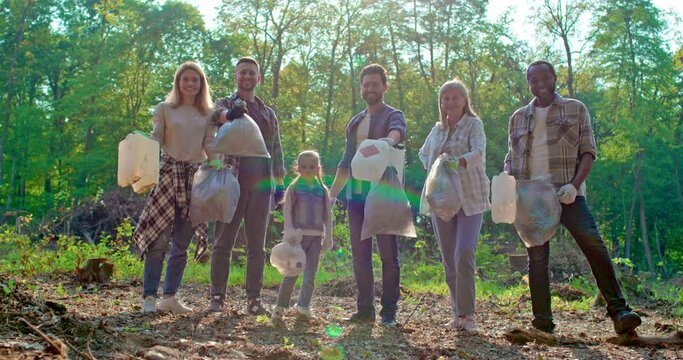 Portrait of multicultural volunteer group standing together in protective gloves and holding garbage bags smiling at camera outdoors. Concept of volunteering, nature protection and charity work.