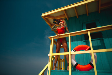 Lifeguard woman in red bikini and visor with binocular and lifebuoy on watch tower against turquoise sky - obrazy, fototapety, plakaty