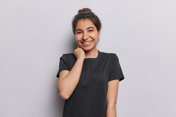 Horizontal shot of pretty Iranian girl with dark hair gathered in bun keeps hand on neck poses happy in studio dressed in casual black t shirt isolated on white background. People and sincere emotions