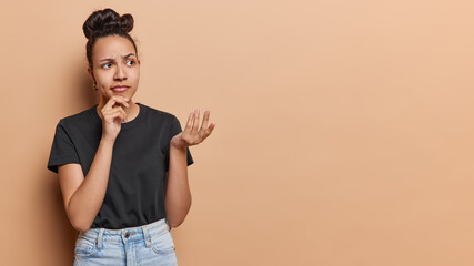 Studio shot of pretty Latin woman with hair bun keeps hand on chin shrugs shoulders with...