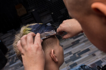 The process of hairdressing services: a professional barber cuts a fashionable young client guy with a comb and scissors in the salon