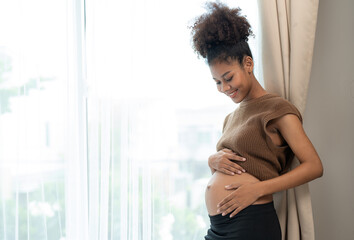Fototapeta na wymiar Happy African American pregnant woman touching belly and standing beside windows in bedroom at home. motherhood, pregnancy, and expectation concept.
