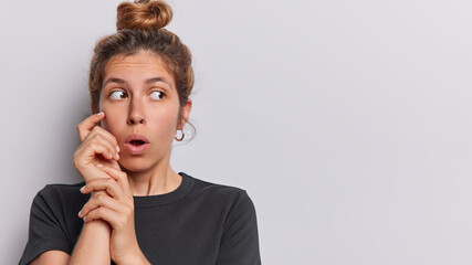 Horizontal shot of shocked young woman with hair gathered in bun looks stunned aside holds breath...