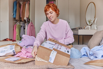 Fototapeta na wymiar Indoor shot of redhead woman prepares delivery box sells her second hand clothes poses on bed against bedroom interior smiles pleasantly. Preparing parcels for shipment. Online marketing concept