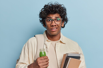 Indoor portrait of young disappointed hindu man isolated on blue background wearing white tshirt and shirt with long sleeves holding white bag on his left shoulder with small glass bottle of smoothie