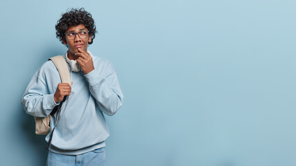 Studio shot of isolated on left side of blue background young thoughtful hindu man with short black curly hair wearing blue jeans and sweater with white big bag on left shoulder and headphones on neck