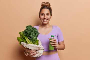 Indoor shot of pleased young sporty woman with hair bun dressed in purple tracksuit poses with fresh smoothie and bag of green vegetables leads healthy lifestyle. Proper nutrition and sport.