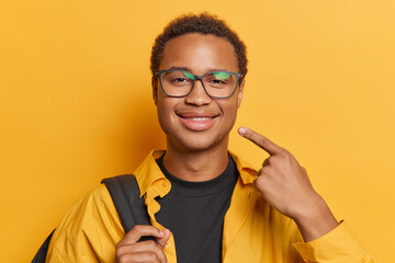 Portrait of handsome dark skinned adult man with short hair points at his smile wears transparent eyeglasses and shirt carries rucksack isolated over yellow background. Look at my charming smile