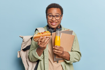 Indoor shot of positive dark skinned man eats hot dog holds bottle of fresh juice carries notepads carries rucksack smiles pleasantly dressed in casual clothes isolated over blue background.
