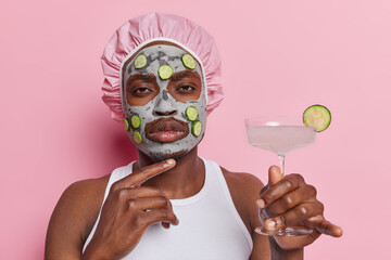Photo of serious dark skinned keeps hand under chin looks directly at camera applies beauty clay mask with cucumber slices holds fresh cocktail isolated on pink background. Beauty and wellness concept
