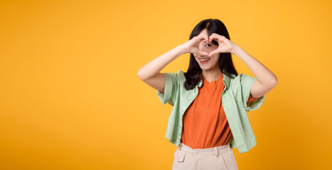 Asian young woman 30s wearing green and orange shirt. With a heart hand gesture on eye, adds whimsical touch. Unique and creative expression of affection on a vibrant yellow background.
