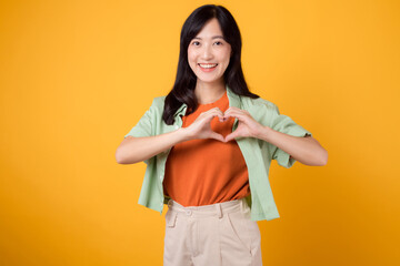 Close up of Asian woman 30s wearing orange shirt showing heart hand gesture, symbolizing affection and warmth. Perfect for expressing love and spreading positive vibes in various projects.
