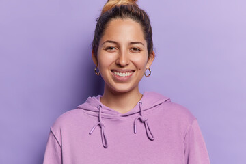 Portrait of cheerful young woman with combed hair smiles toothily expresses happy emotions shows white teeth dressed in casual sweatshirt isolated over purple background. People and happiness concept