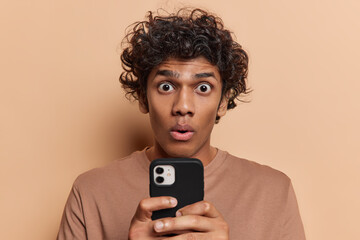Horizontal shot of young hindu man wearing brown tshirt with black curly short hair holding black smartphone in front of him looking little bit shocked reading news isolated on beige background