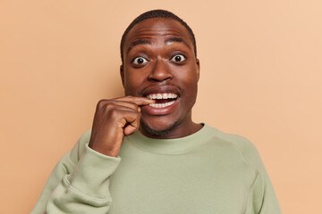 Photo of surprised dark skinned man bites finger looks surprisingly at camera notices something has curious expression wears casual jumper isolated over brown background notices something breathtaking