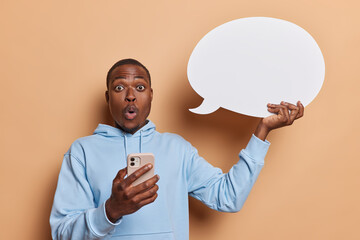 Shocked dark skinned man holds smartphone and white speech bubble suggests to write your promotional content dressed in casual blue sweatshirt isolated over beige background. Technology concept