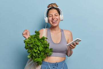 Joyful pleased European sportswoman dressed in cropped top and shorts listens mucis via headphones connected to smartphone carries fabric bag with green parsley isolated over blue background