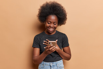 Smiling happy attractive young african woman wearing blue jeans and grey tshirt holds mobile phone at her hand. She is delighted receiving message with good news. Isolated on brown background