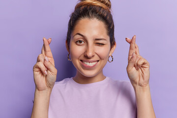 Photo of hopeful young woman radiates joy as she crosses fingers anticipating luck winks eyes and smiles broadly dressed in casual t shirt isolated over purple background. Hope and good fortune