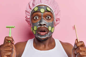 Horizontal shot of impressed dark skinned man applies facial clay mask and cucumber slices keeps mouth opened holds razor and toothbrush undergoes hygiene routines isolated over pink background