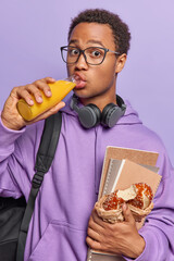 Vertical image of surprised curly African schoolboy drinks fresh juice holds notepads and delicious bun wears transparent eyeglasses and hoodie carries rucksack isolated over purple background