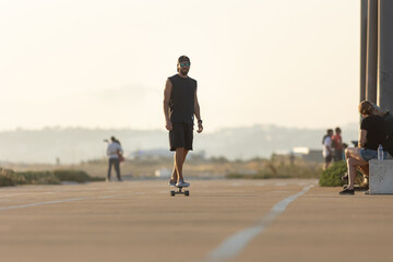 Adult attractive smiling man in black clothes skating on the street by the beach