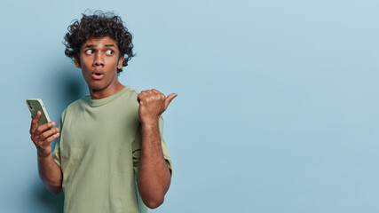 Waist up shot of curly Hindu man points thumb away on copy space holds mobile phone demonstrates something awesome dressed in green t shirt isolated over blue background. People and technology concept