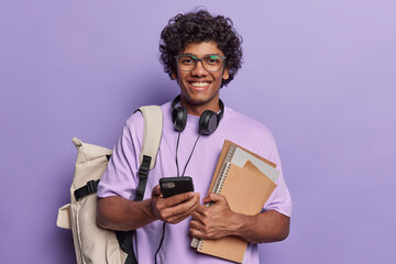 Horizontal shot of pleased curly haired Hindu student smiles gladfully uses modern texhnologies for studying and entertainment carries notepads carries backpack wears t shirt and spectacles.