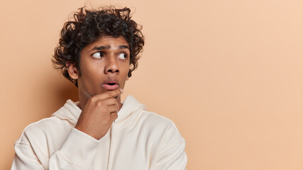 Studio shot of young thoughtful hindu guy with black short curly hair and slightly open mouth wearing white hoodie holding one of his hands on chin turning head to right side trying to solve problem
