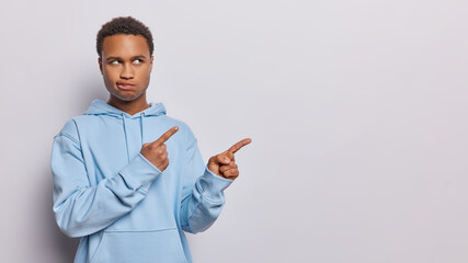 Horizontal shot of serious displeased dark skinned African man purses lips has angry expression points with both index fingers at empty space wears blue hoodie isolated over white background.