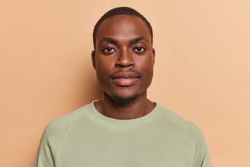 Portrait of serious handsome dark skinned man looks directly at camera with concentrated gaze dressed in casual jumper poses in studio against brown background for making photo on documents.