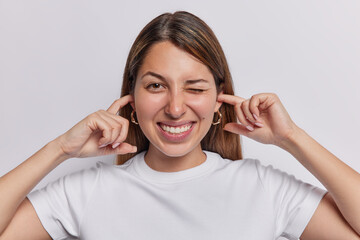 Photo of lovely young woman with long hair pretending not to hear what she is told plugs ears winks eye shows teeth dressed in casual t shirt isolated over white background. Body language concept
