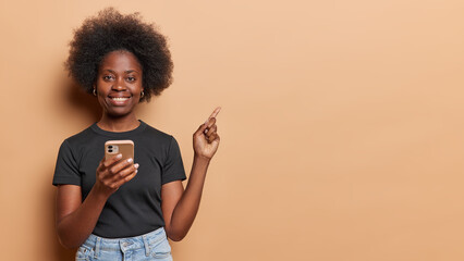 Young beautiful smiling cheerful african woman wearing grey nice tshirt and blue jeans holding her mobile phone at hand trying to make selfie holding one of fingers up pointing at something.