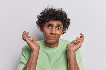 Photo of hesitant curly haired man spreads palms feels unaware what to do dressed in green basic t shirt cannot make decision isolated over white background. Human perception and reactions concept
