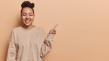 Happy teenage girl with dark hair dimples on cheeks stands with closed eyes smiles broadly dressed in casual jumper points index finger on copy space for your promotion isolated over brown background
