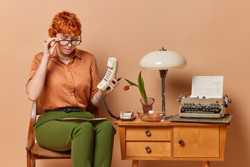 Attentive busy redhead young businesswoman or secretary wears spectacles concentrated at paper document holds handset studies financial report isolated over brown background. Retro style concept
