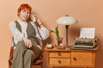 Horizontal shot of ginger young female model dressed elegantly has telephone conversation via...