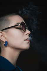 Close up portrait of short hair female wearing round sunglasses. Exhailing thick sigarette smoke. Dark ambient environment.